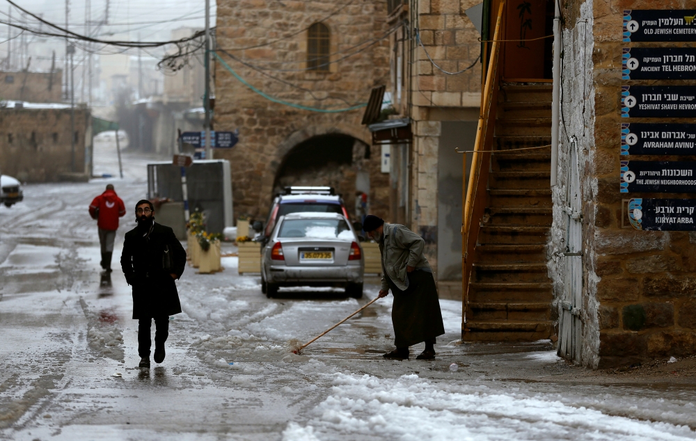 A Jewish settler walks as a Palestinian man removes snow during a winter storm in Hebron, in the Israeli-occupied West Bank, on Thursday. — Reuters