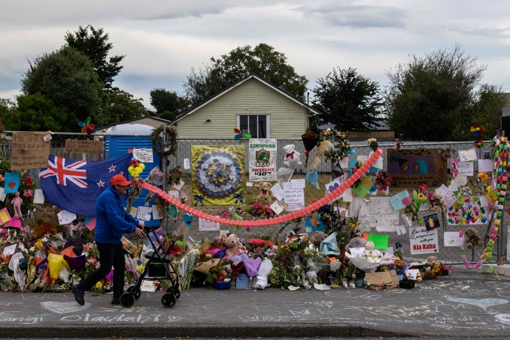 A man man walks past flowers and tributes displayed in memory of the twin mosque massacre victims along the road outside the Linwood mosque, back center, in Christchurch, New Zealand, on Monday. — AFP