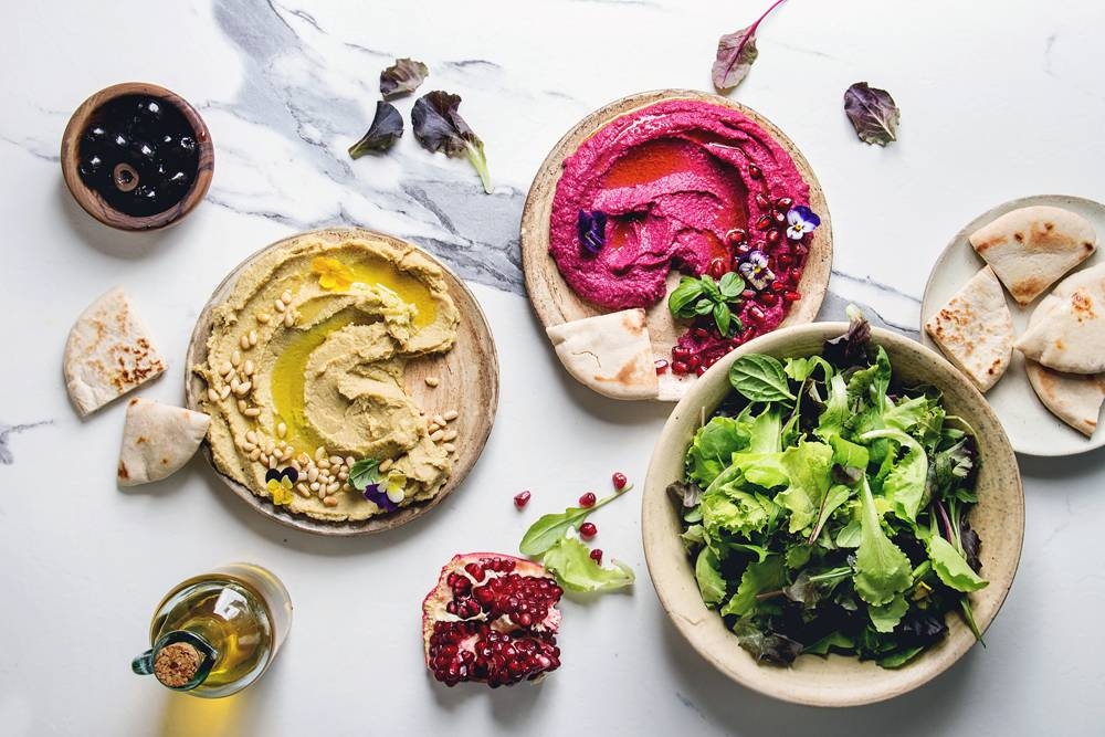 Variety of homemade traditional and beetroot spread hummus with pine nuts, olive oil, pomegranate served on ceramic plates with pita bread and green salad on white marble background.