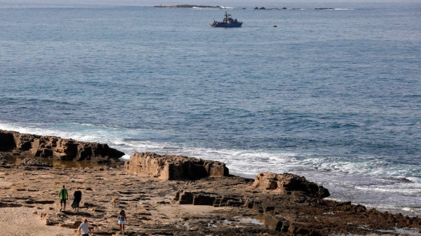 People walk along the beach as a navy vessel patrols the Mediterranean waters Ras al-Naqura on the Israeli side of the border between the two countries.