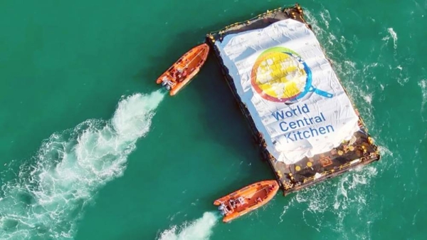 Orange dinghies approach the aid barge. There is a sheet over the aid that reads 