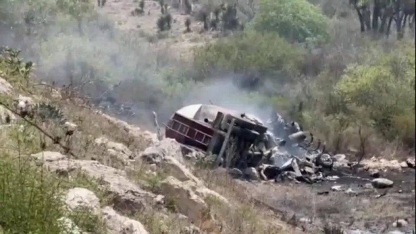 The wreckage of a vehicle surrounded by smoke at the bottom of a rocky hill