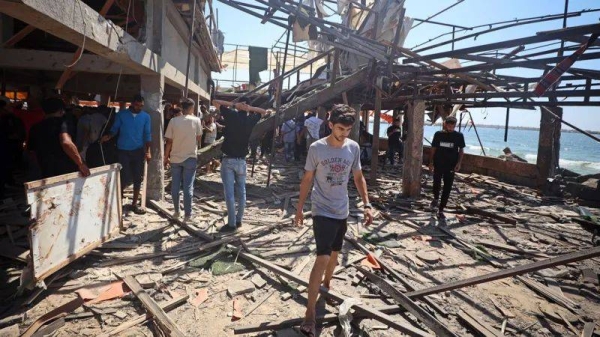 A view of the destruction after air strikes on a coffee shop in Gaza. People are pictured examining the debris with a young man walking out of the cafe in the foreground, and the sea and blue sky in the background, taken on 30 Jun