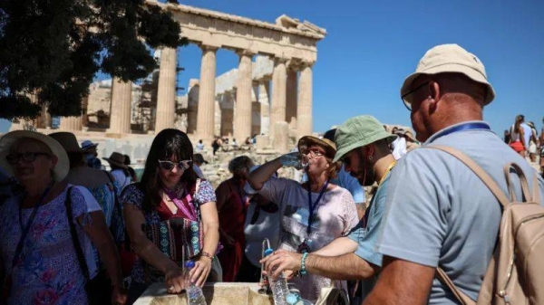 A small group of men and women wearing sunhats fill up their water bottles near a shaded area in front of the Acropolis  with a crowd of visitors in the background