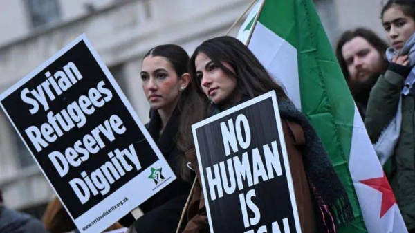 Two female protesters hold Syrian flags and placards reading, 'Syrian refugees deserve dignity' and 'no human is illegal'