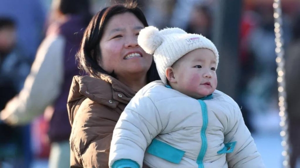 A woman, in a beige coat, holding a baby, who is wearing a white coat with turquoise trim and a white woollen hat, in a park in Fuyang, east China's Anhui province. Photo taken on 16 January, 2025.