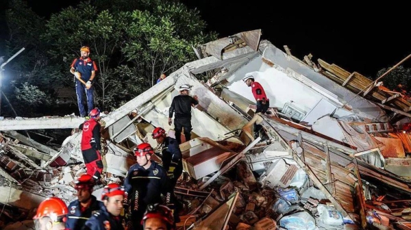 Emergency workers in blue and red uniforms stand on a building that has crumbled from the earthquake. It is night time and the debris is light brightly by spot lights